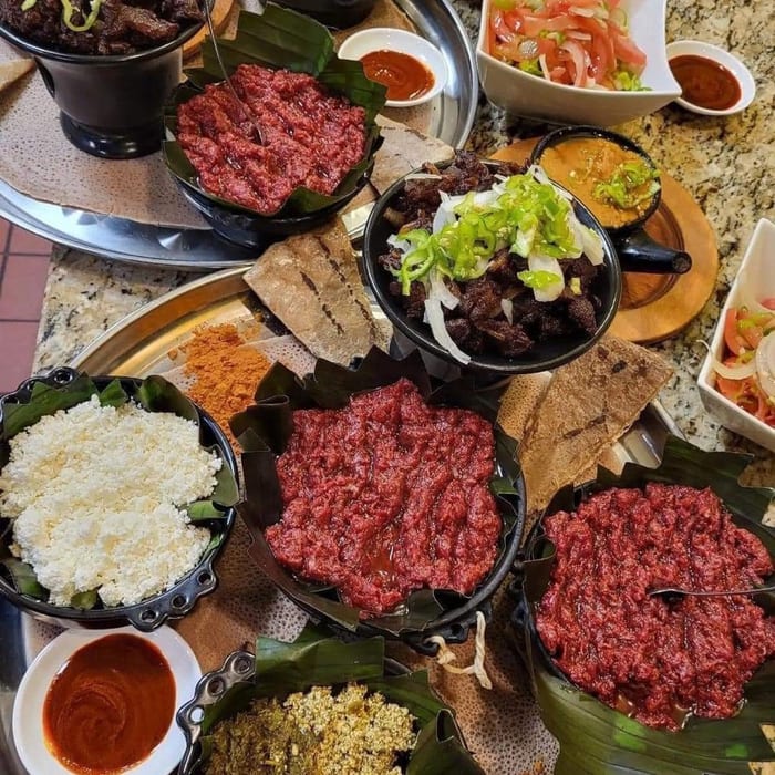 A large catering spread of kitfo, ayib, gored gored, tibs and greens in banana-leaf lined dishes on injera trays