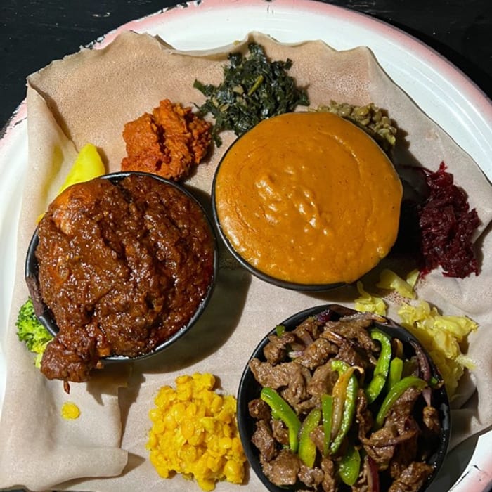 A plate of injera topped with key wat, shiro, gomen, tibs with green peppers, kik alicha and beetroot